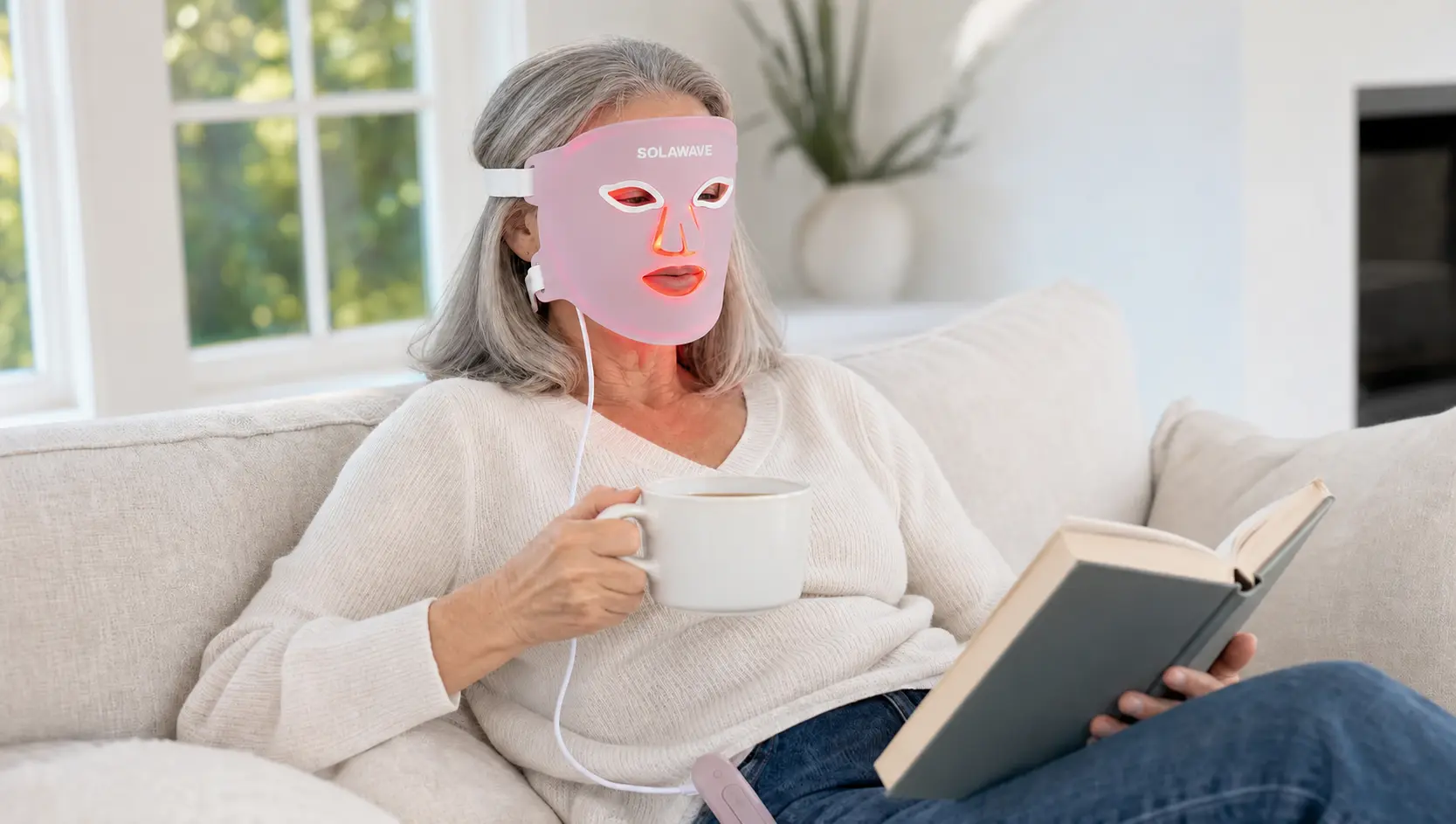 A silver-haired woman wearing a Solawave mask on a sofa with a book and mug; editorial illustration
